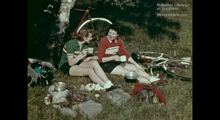 Two women are taking a break in the countryside. They are wearing sporting clothes and drinking from cups. There is a bicycle and a tree behind them.