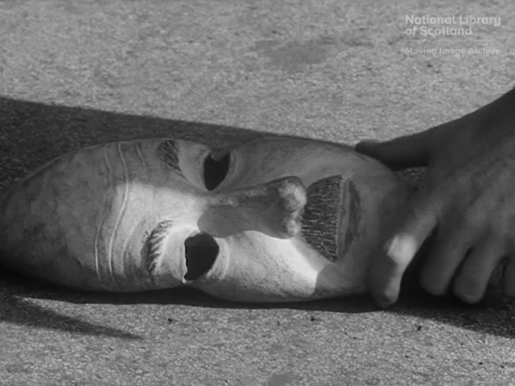 Black and white still of rubber mask on ground with a man's hand holding it