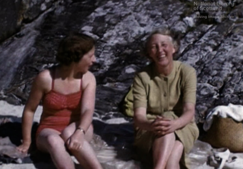 film still in colour of beach. Two white women sitting site by side. One on right wearing red bathing suit. Older woman beside wearing green dress. There is a picnic hamper.