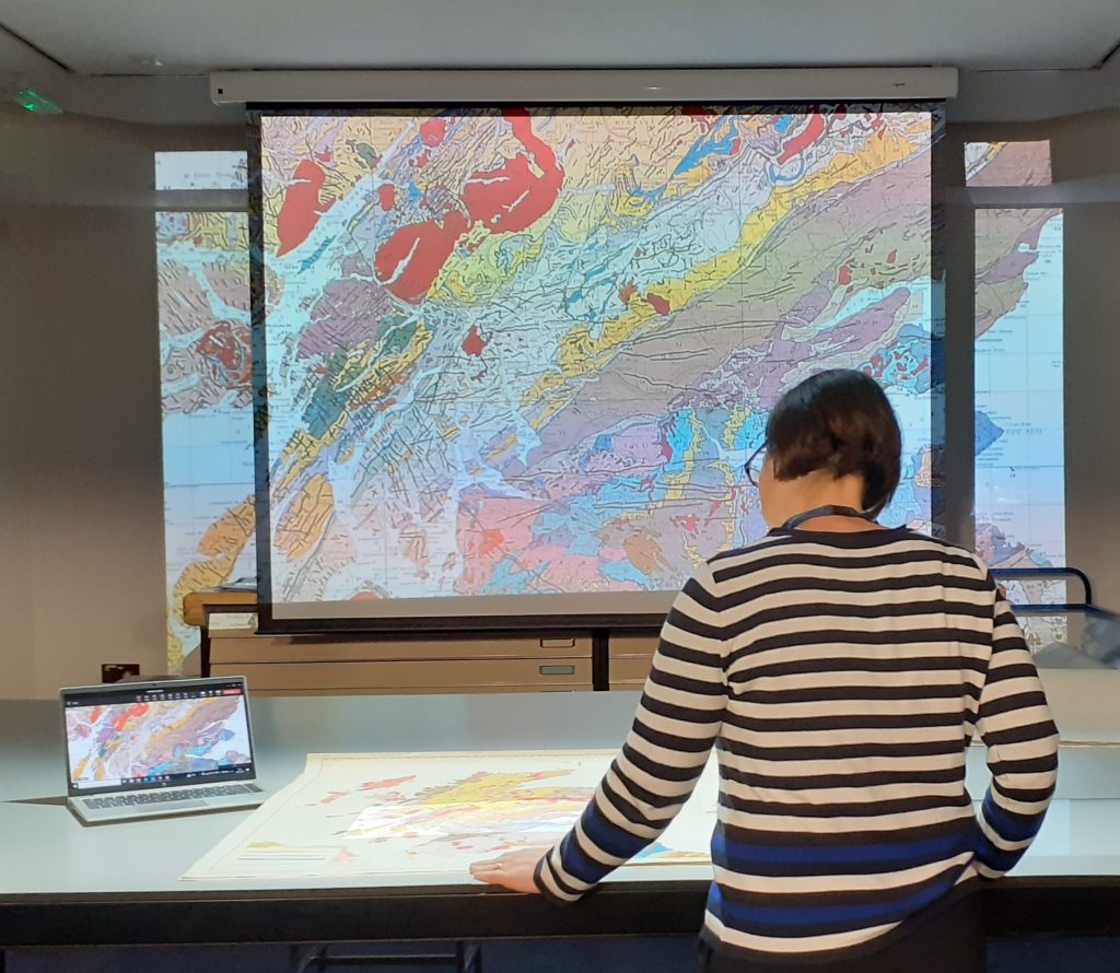 Image of the ceiling visualiser being set up. There is a map on the table with a member of staff looking at it. Just ahead is a projected image of the map on a large screen. To the left of the staff member is a laptop showing the map.