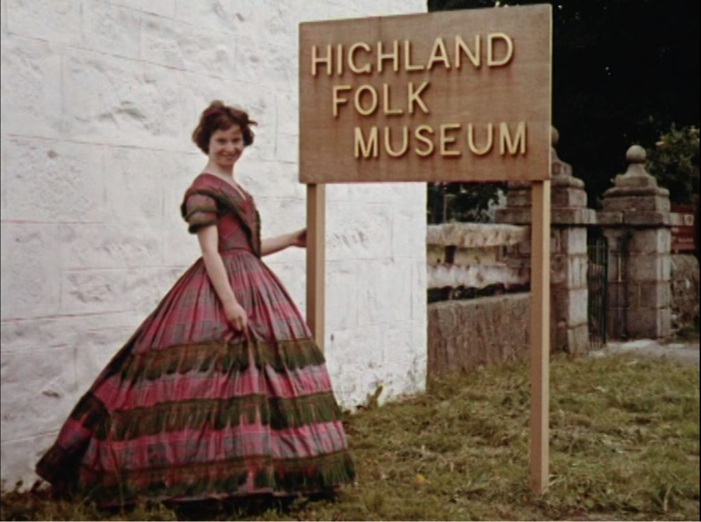 A woman in Highland dress stands at sign for the Highland Folk Museum 